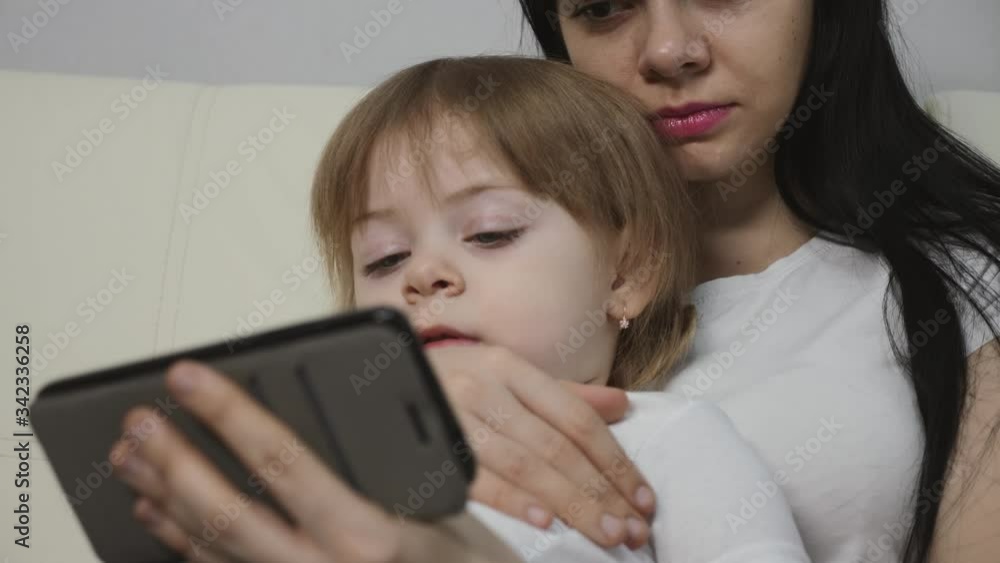 A dark-haired young mother and her little blonde daughter are sitting on the couch at home and using a smartphone to learn and communicate. A dark-haired young mother and her little blonde daughter are sitting on the couch at home and using a smartphone to learn and communicate.