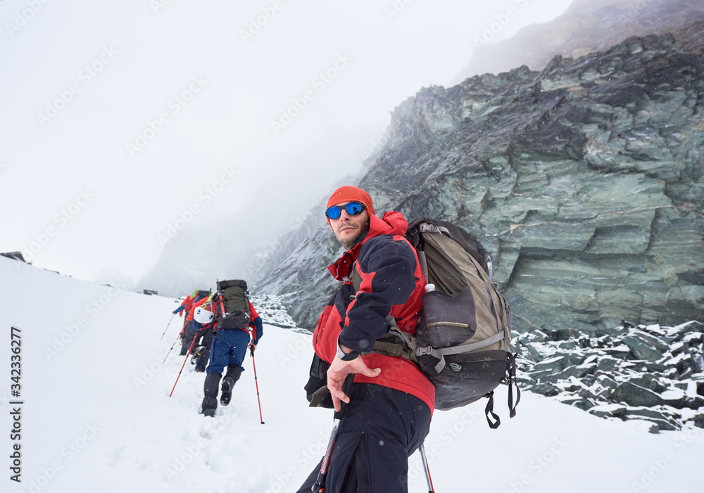 Side view of hiker in sunglasses looking at camera while friends walking on mountain path covered with snow. Male traveler with backpack standing in winter valley with big rock on background. Side view of hiker in sunglasses looking at camera while friends walking on mountain path covered with snow. Male traveler with backpack standing in winter valley with big rock on background.