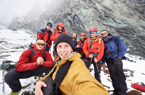 Cheerful young man taking picture with friends hikers during travel in winter mountains. Group of male travelers looking at camera and smiling. Concept of travelling, hiking and photography. photo