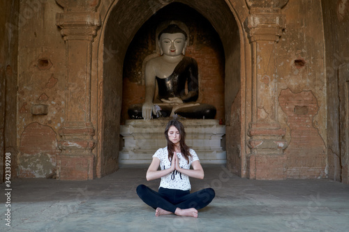 A beautiful girl is meditating in lotus position, sitting on the floor in front of a Buddha statue in an ancient temple in Bagan in Myanmar.