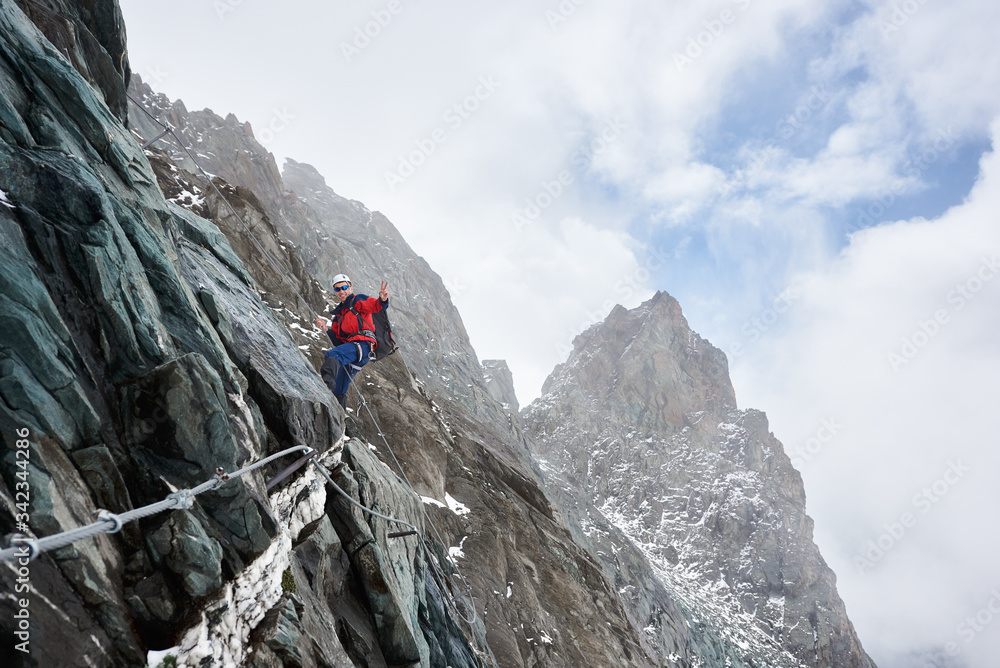 Side view of male alpinist in sunglasses and safety helmet holding fixed rope while ascending mountain. Mountaineer showing victory gesture and looking at camera with smile. Concept of mountaineering