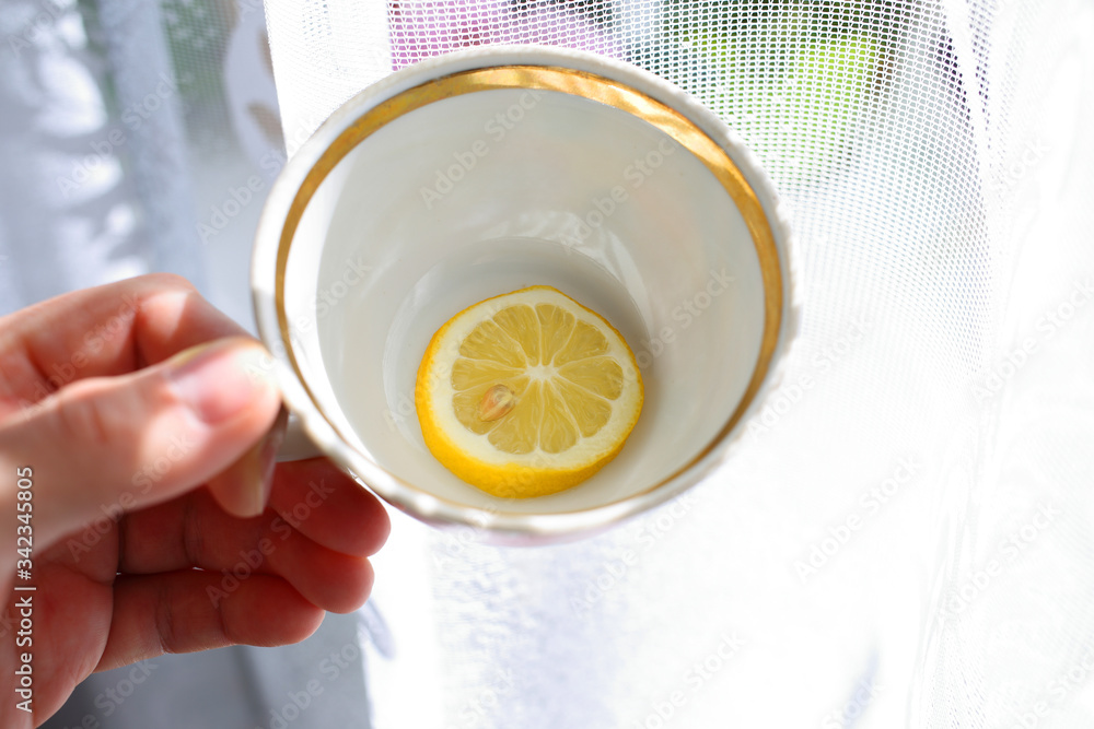 Caucasian man's hand holds an empty white mug with gold border with round slice of juicy yellow fresh lemon with grain at bottom, against background of perforated fabric backlit by sunlight Caucasian man's hand holds an empty white mug with gold border with round slice of juicy yellow fresh lemon with grain at bottom, against background of perforated fabric backlit by sunlight