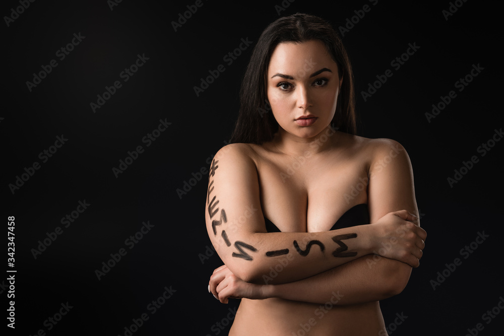 Fototapeta premium Front view of attractive plus size model with lettering hashtag feminism on body covering bust with hands isolated on black