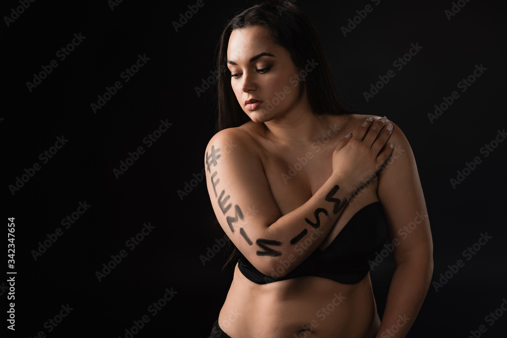 Front view of plus size model with lettering hashtag feminism on body covering breast with hand isolated on black Front view of plus size model with lettering hashtag feminism on body covering breast with hand isolated on black