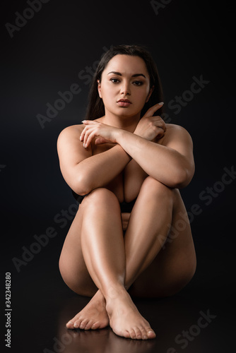 Front view of beautiful plus size girl sitting with crossed legs and hands on black background