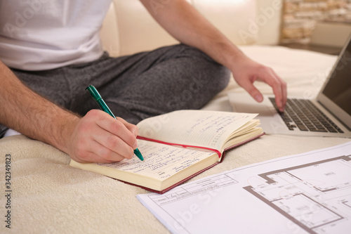 Unrecognizable male hands of a businessman on self-isolation during the COVID-19 coronavirus pandemic, works at home, sits on the bed, does his work remotely, notepad, documents and laptop close up