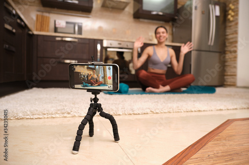 Athletic woman blogger in sportswear shoots video on phone as she does exercises at home in the kitchen. Stay at home activities. Prevent infection from the pandemia.