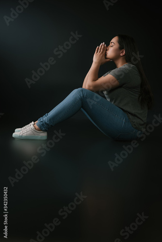 Side view of upset plus size girl covering face with hands and sitting on black background
