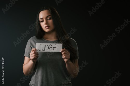 Front view of sad plus size girl holding card with word judge on black background