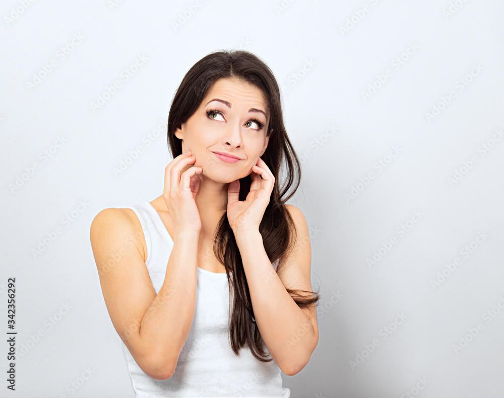 Beautiful curious smiling woman holding the head the hands thinking and looking up in casual white t-shirt and long hair on blue background. Closeup Beautiful curious smiling woman holding the head the hands thinking and looking up in casual white t-shirt and long hair on blue background. Closeup