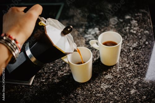 Preparing fresh coffee in moka pot on electric stove. Pouring hot steaming coffee into two white mugs.