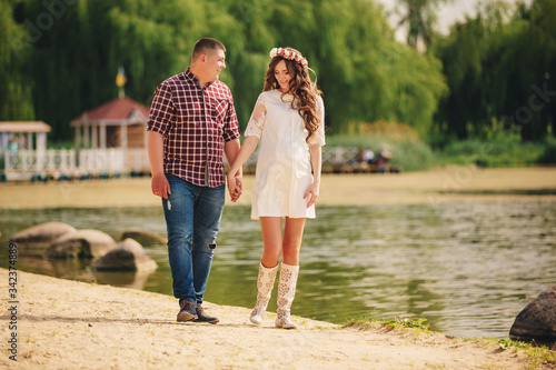 Young happy romantic pregnant couple are walking on nature near lake in summer park. Pregnant woman expecting a baby. Future mom and dad, family. mother's, father's day