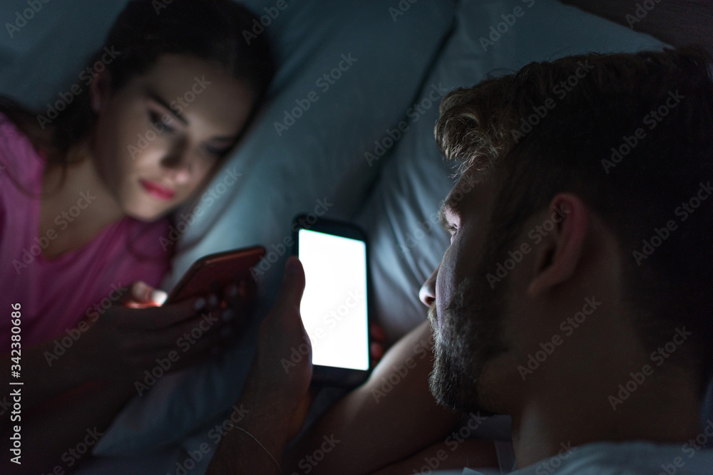 Selective focus of man using smartphone near girlfriend chatting on bed at night Selective focus of man using smartphone near girlfriend chatting on bed at night