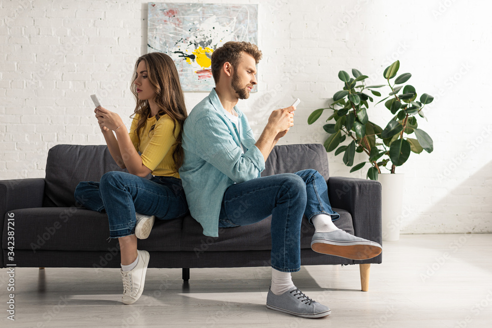 Fototapeta premium Side view of couple using smartphones while sitting back to back on sofa