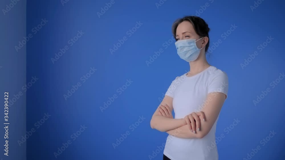 Portrait of woman wearing medical face mask, white shirt, looking away in room with blue wall at home: slow motion. Self isolation, prevention, quarantine, COVID19, coronavirus, safety concept Portrait of woman wearing medical face mask, white shirt, looking away in room with blue wall at home: slow motion. Self isolation, prevention, quarantine, COVID19, coronavirus, safety concept
