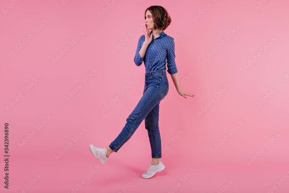 Full-length photo of shapely glad girl in white sneakers. Slim debonair young woman in jeans posing on pastel background. Full-length photo of shapely glad girl in white sneakers. Slim debonair young woman in jeans posing on pastel background.