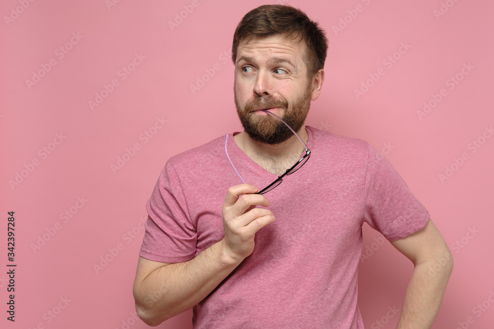 Doubting, bearded man in a pink tee-shirt suspects someone, he holds glasses in mouth and looks oddly in the side. Doubting, bearded man in a pink tee-shirt suspects someone, he holds glasses in mouth and looks oddly in the side.