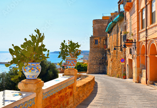 Old narrow street with Ceramic pot with cactus on wall and view on the port and sea in front of Sciacca, Sicily, Italy