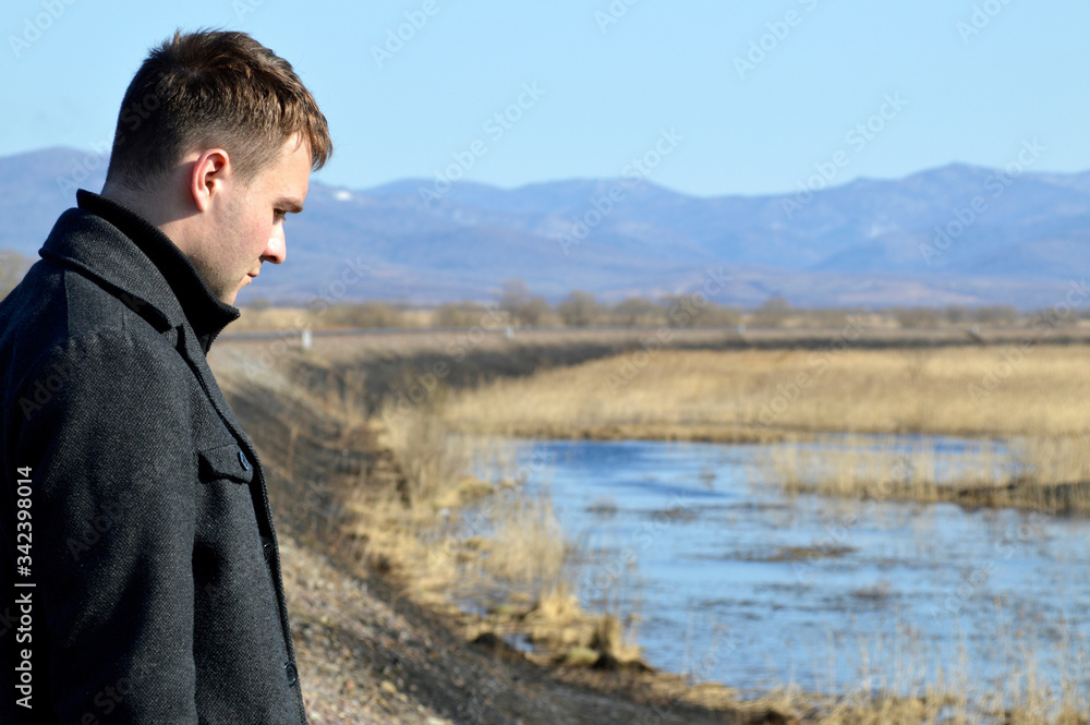 Young man in coat looking down with upset face near swamp and rail. Depressive concept loss of money, loneliness, unemployment. Copy space Young man in coat looking down with upset face near swamp and rail. Depressive concept loss of money, loneliness, unemployment. Copy space