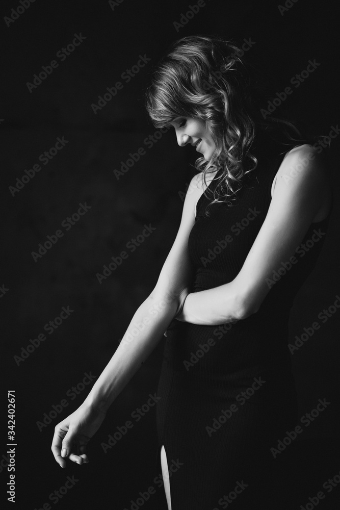Beautiful elegant slim woman in a black dress in a loft studio modestly looks down. Soft selective focus. Black and white art photo.