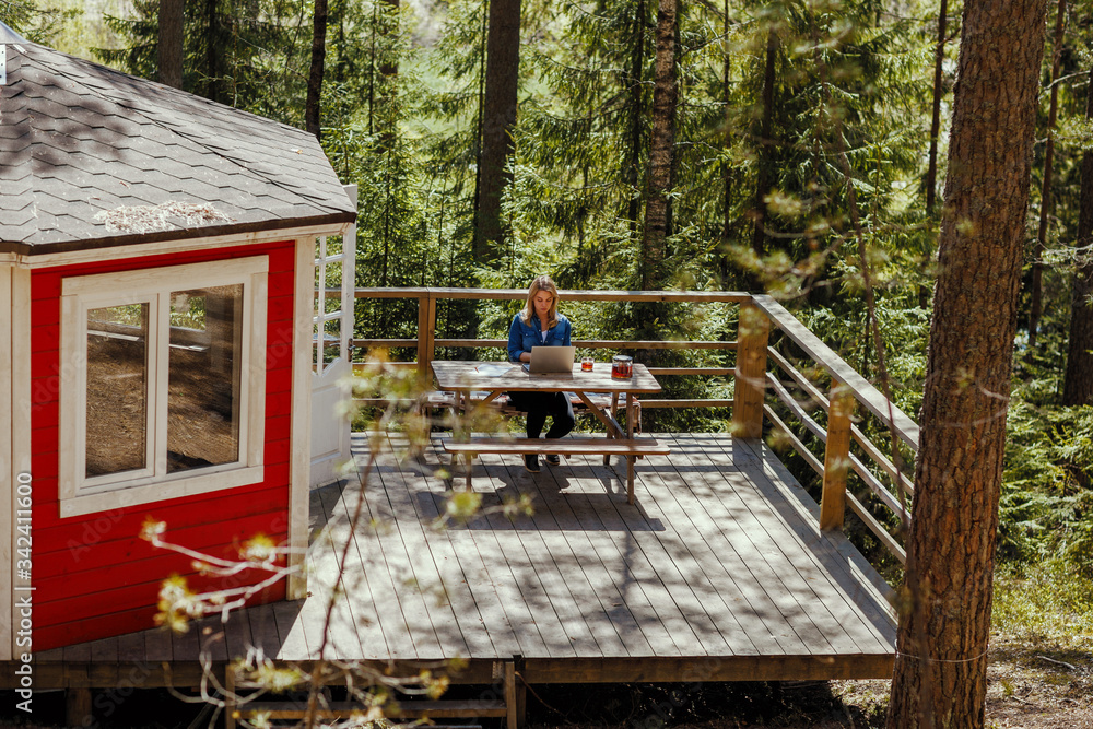Young woman working remotely on laptop sitting at table on wooden terrace of country house surrounded by pine trees