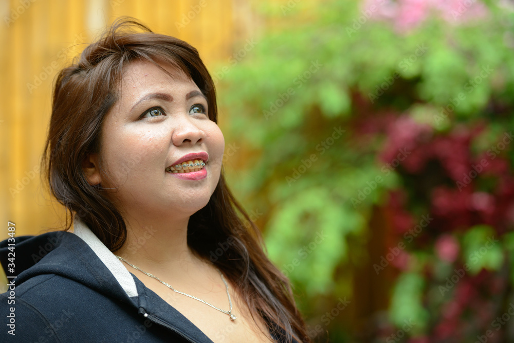 Face of happy young overweight Asian woman thinking outdoors Face of happy young overweight Asian woman thinking outdoors