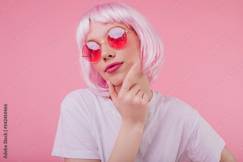 Pensive charming girl with pink hair looking up while posing on pastel background. Carefree european woman in sunglasses and periwig chilling during studio photoshoot. Pensive charming girl with pink hair looking up while posing on pastel background. Carefree european woman in sunglasses and periwig chilling during studio photoshoot.