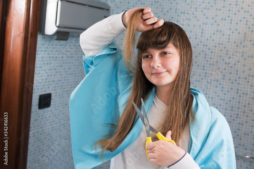 A beautiful young girl is holding a scissor to cut her hair herself during lock down of coronavirus, looking in mirror. DIY hair cut during the epidemic of covid-19, stay at home, selective focus