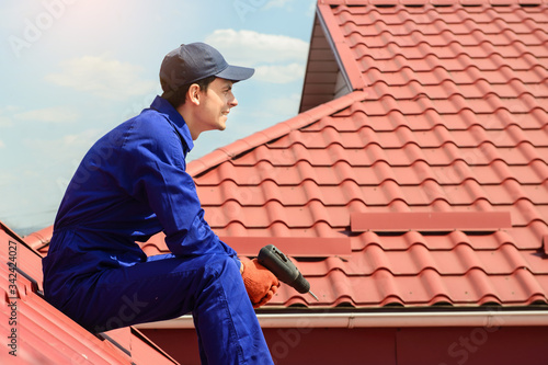 Young happy man contractor worker in blue overalls is repairing a red roof with electric screw driver and have a rest on the roof