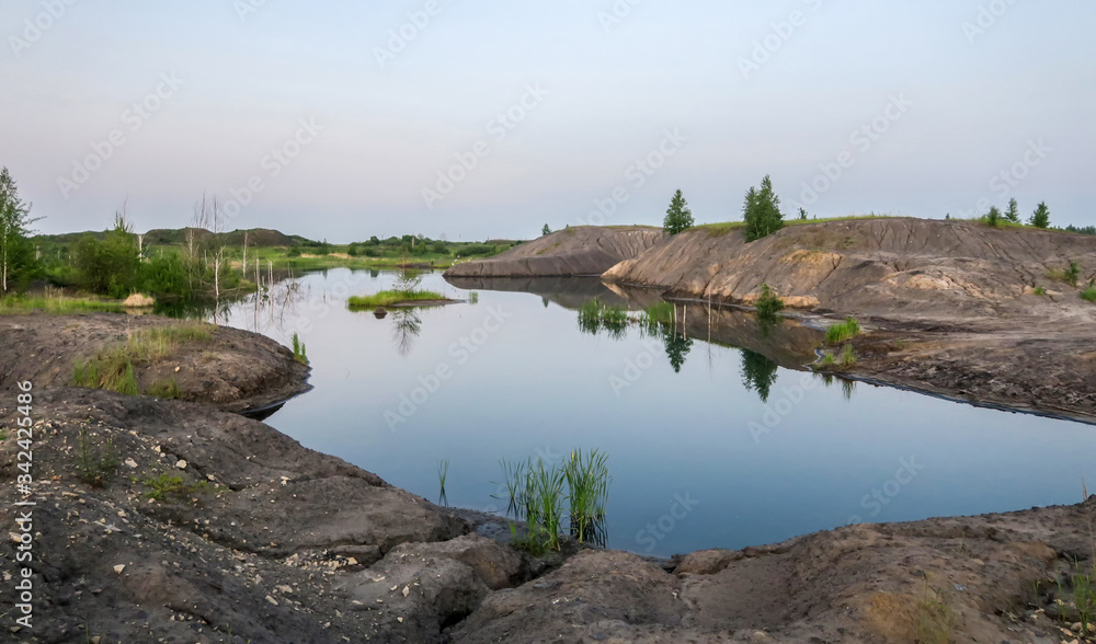 Lake in a coal pit with clear water at sunset. Reflection in the water of a coal mountain, hill, trees, reeds.The fuel industry produces coal in an open way.
