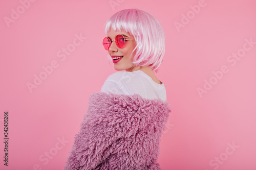 Portrait of playful girl in shiny pink peruke and sunglasses. Debonair caucasian woman looking over her shoulder with smile.