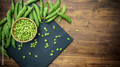 Top down flat lay view of fresh shelled peas on a black slate plate and some pods on a wooden background. Black, white and green copy space composition for healthy vegan cuisine