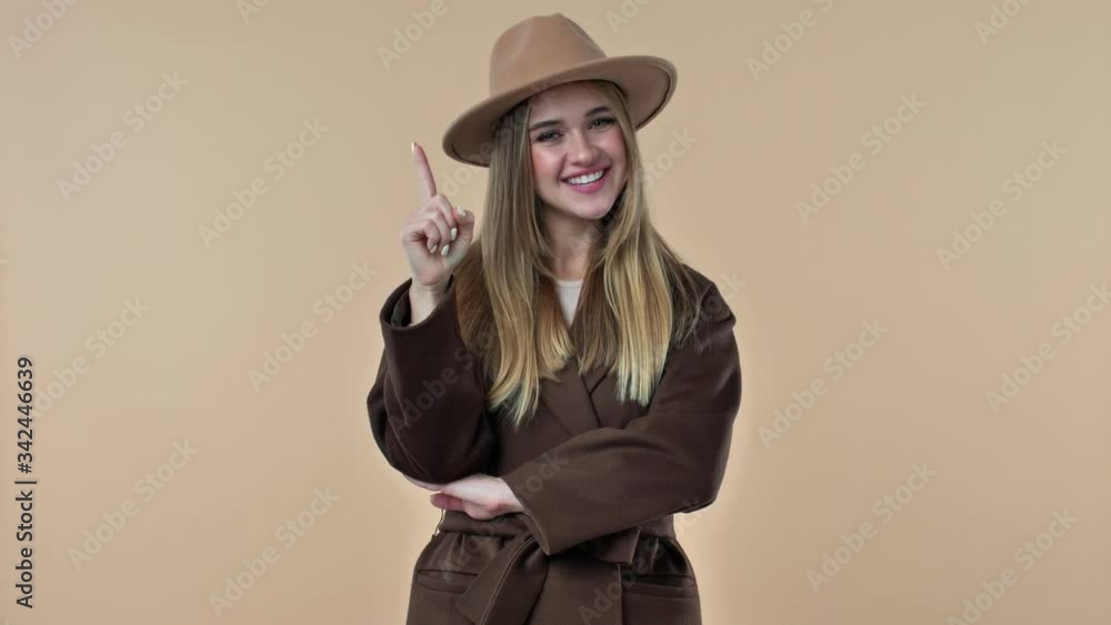 A thoughtful young woman wearing hat and coat is coming up with the idea over the beige background in studio A thoughtful young woman wearing hat and coat is coming up with the idea over the beige background in studio