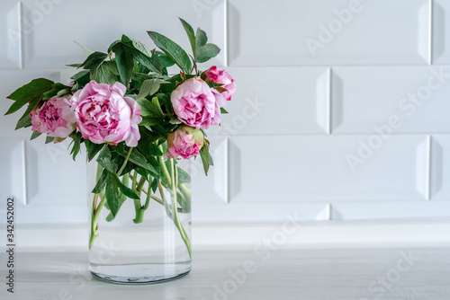 Bouquet of pink peonies in glass vase against white background and wooden table. Mothers day card. Beautiful peony flower for catalog or online store. Floral shop and delivery concept. Copy space.