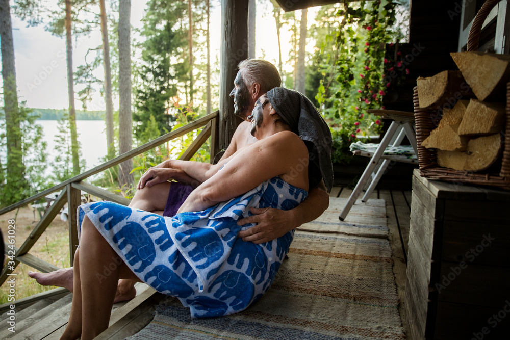 Aged couple having black facial mask after Finnish sauna. Mature man and woman in towels sitting on wooden steps, relaxing, hugging and enjoying the lake view. 