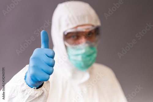 Male doctor with surgical mask, goggles and protective suit pointing okay on gray backround.