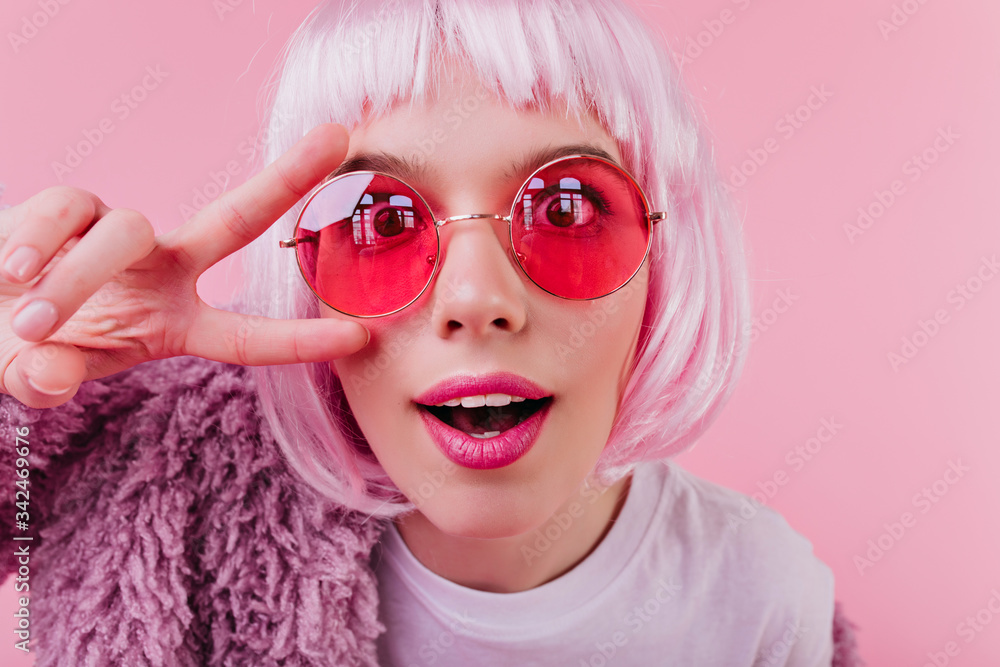 Close-up shot of funny european girl in colorful periwig and pink sunglasses. Indoor photo of attractive lady in short peruke fooling around in studio.