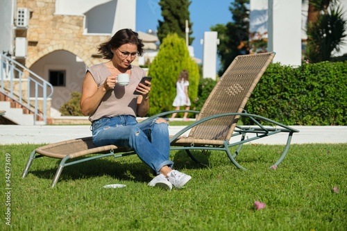 Beautiful woman resting sitting on sunbed on green lawn, reading smartphone © Valerii Honcharuk