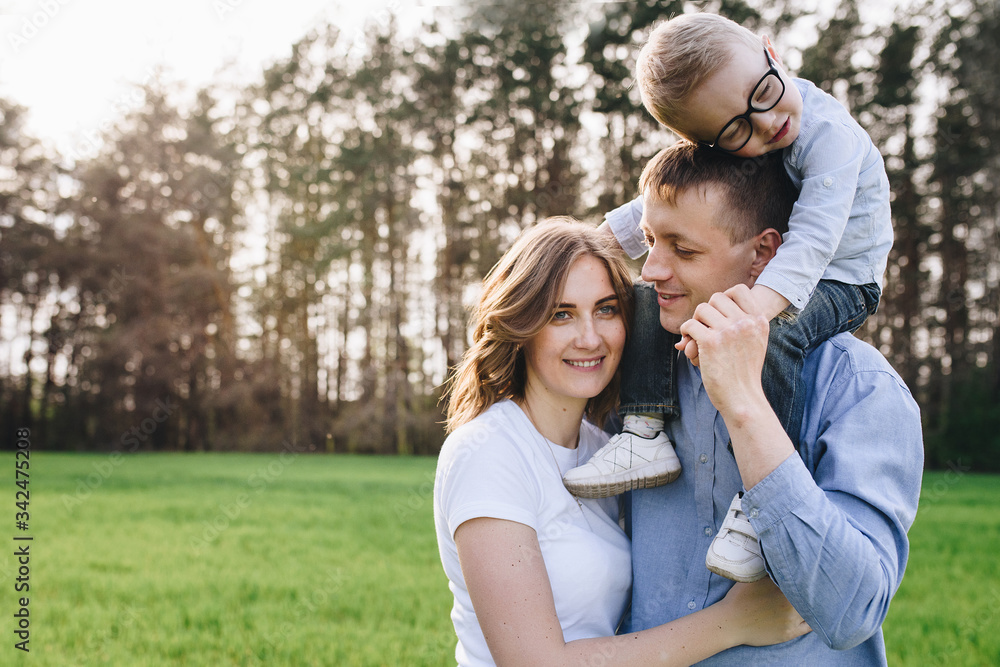 Family in the forest at a picnic. Sit in a clearing, green grass. Blue clothes. Mom and Dad play with their son, hug and smile. A child with glasses.