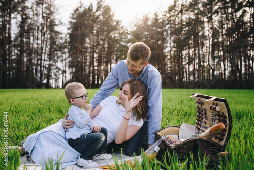 Family in the forest at a picnic. Sit in a clearing, green grass. Blue clothes. Mom and Dad play with their son, hug and smile. A child with glasses.