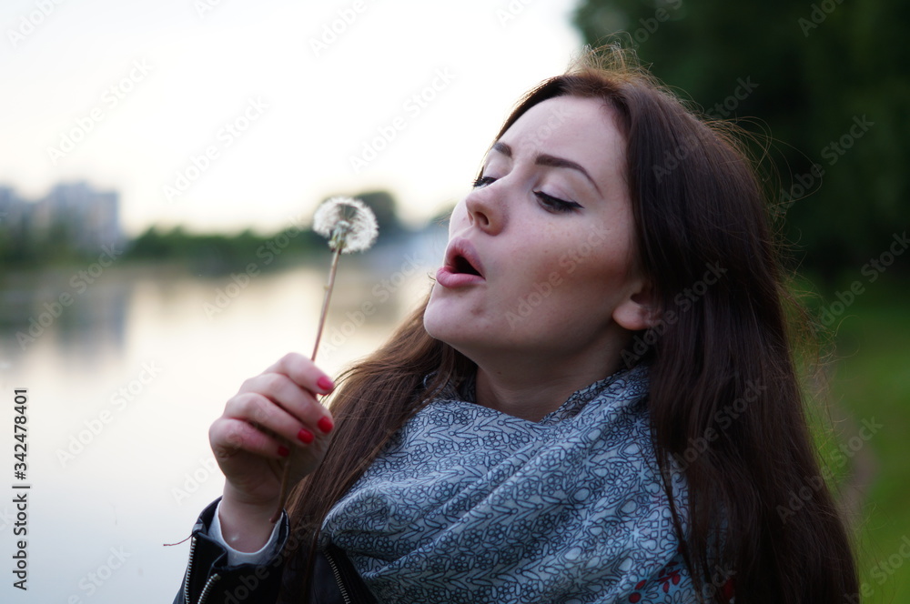 A beautiful girl blows in a dandelion and its fluff scatters in nature. It's summer outside. Woman in a scarf and a black jacket with long hair A beautiful girl blows in a dandelion and its fluff scatters in nature. It's summer outside. Woman in a scarf and a black jacket with long hair