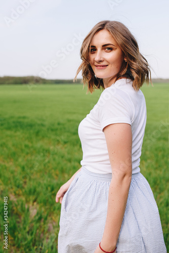 The girl is blonde, brown hair, in a white shirt and blue midi skirt. Walking in the field, through the green grass. Portrait of a girl. Positive and smile.