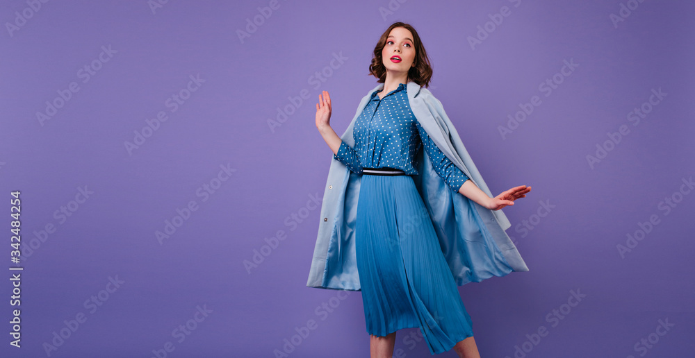 Emotional brunette woman in blue coat posing on purple background in studio. Indoor photo of beautiful short-haired female model in trendy midi dress. Emotional brunette woman in blue coat posing on purple background in studio. Indoor photo of beautiful short-haired female model in trendy midi dress.