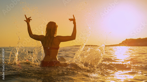 LENS FLARE Young woman sitting on a surfboard playfully splashes water at sunset