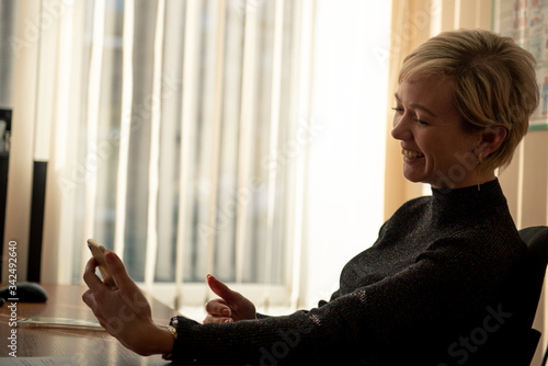 A young woman with a short haircut looks at the phone screen and laughs while sitting in an office chair at her desk