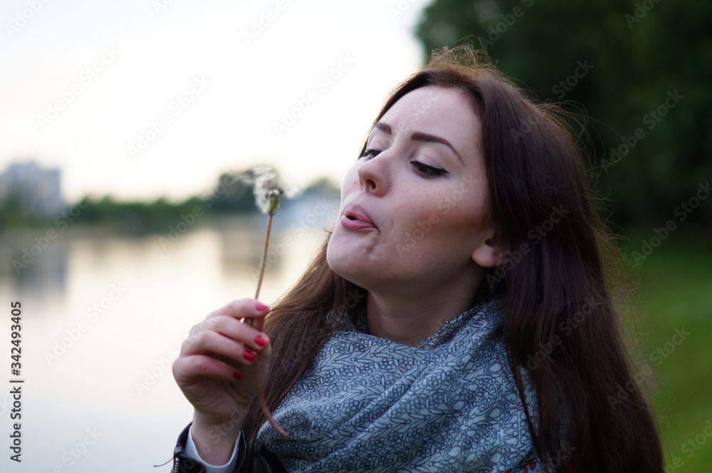 A beautiful girl blows in a dandelion and its fluff scatters in nature. It's summer outside. Woman in a scarf and a black jacket with long hair A beautiful girl blows in a dandelion and its fluff scatters in nature. It's summer outside. Woman in a scarf and a black jacket with long hair