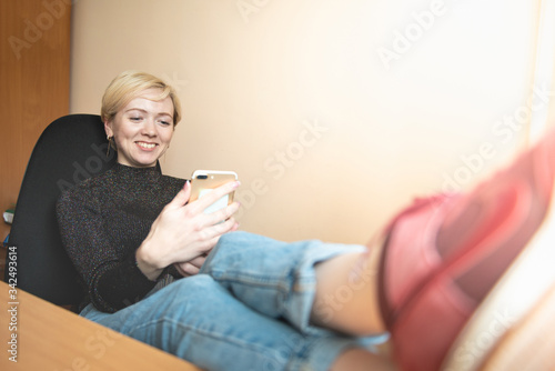 A young woman with a short haircut looks at the phone screen and laughs while sitting in an office chair and putting her legs on the table
