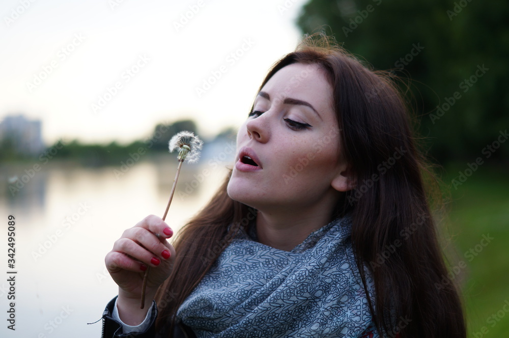 A beautiful girl blows in a dandelion and its fluff scatters in nature. It's summer outside. Woman in a scarf and a black jacket with long hair A beautiful girl blows in a dandelion and its fluff scatters in nature. It's summer outside. Woman in a scarf and a black jacket with long hair