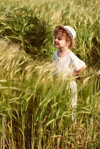 A cute beautiful caucasian boy dressed in a linen cloth and white hat standing in a field of green wheet