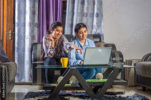 two girls sitting in a living room using laptop and books for online classes